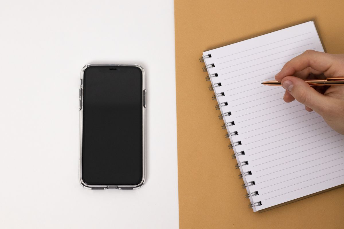 A hand takes notes in a notebook beside a phone.