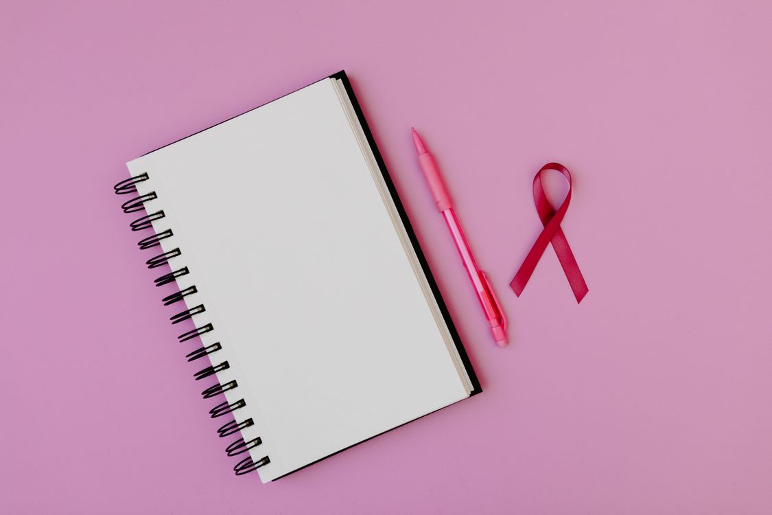 An open notebook, a pen and a pink ribbon on a pink background.